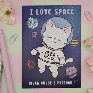 May include: A whimsical illustration features a white cat astronaut with a blue space helmet, surrounded by cat-shaped planets against a starry background. The text "I LOVE SPACE (Both outer & personal)" is displayed. Pink and green accents surround the image.