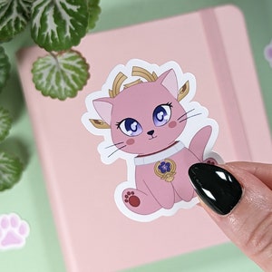 May include: A pink cat sticker with a gold crown and a flower on its chest. The cat has blue eyes and a white outline.