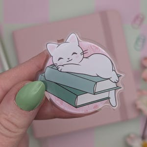 Könnte beinhalten: Ein klarer Katzenaufkleber aus Acryl. Der Aufkleber zeigt eine weiße Katze, die auf zwei türkisfarbenen Büchern liegt. Der Aufkleber hat einen rosa Hintergrund mit einem floralen Muster. Die Katze lächelt. Der Aufkleber wird von einer Hand mit einem grün lackierten Nagel gehalten.