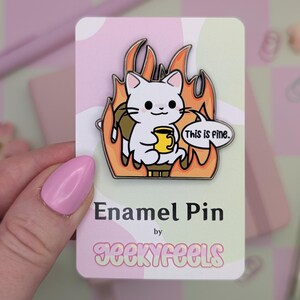 May include: Enamel pin showing a white cat in flames, holding a yellow mug, with the text "This is fine" in a speech bubble. The pin is on a pink and green card with the text "Enamel Pin" and "geekyfeels".