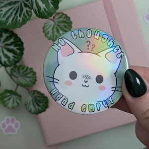Könnte beinhalten: Ein runder Knopf mit einer weißen Katze mit rosa Ohren und einem holografischen Finish. Der Knopf sagt "No thoughts, head empty" mit Fragezeichen über dem Wort "thoughts".