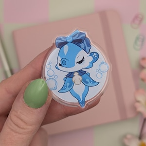 May include: A clear, circular sticker with a cartoon seahorse design. The seahorse has blue details, a bow, and is surrounded by bubbles. The sticker is held in a hand, with a pink notebook and pen visible in the background.