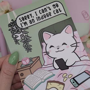 May include: A greeting card with a cartoon cat in bed holding a phone. The card has the text "Sorry, I can't go I'm an indoor cat." The card is light green with pink and white accents, and a small nightstand with a cup of tea.