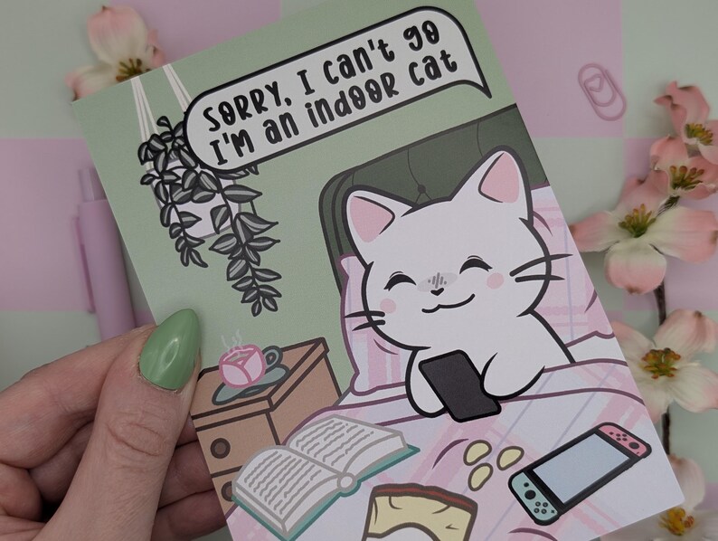 May include: A greeting card with a cartoon cat in bed holding a phone. The card has the text "Sorry, I can't go I'm an indoor cat." The card is light green with pink and white accents, and a small nightstand with a cup of tea.