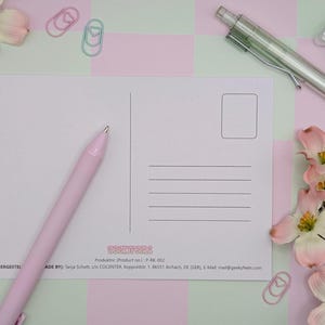 May include: A blank postcard with lines for addressing and a stamp box, alongside a pink pen, a green mechanical pencil, and a light blue eraser. The postcard is surrounded by pink and white flowers, and pink and green paperclips.