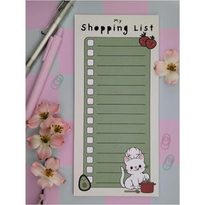 Könnte beinhalten: Notizblock für Einkaufslisten mit dem Text "My Shopping List" oben. Der Notizblock hat einen grünen Hintergrund mit weißem Rand und eine Cartoon-Illustration einer Kochkatze. Rosa Blumen und Stifte im Hintergrund.