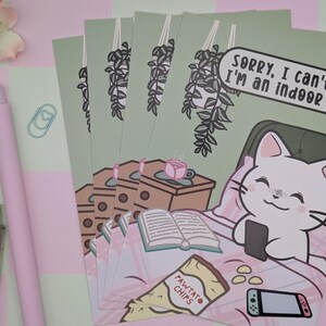 May include: A stack of art prints featuring a cartoon cat in bed with a phone, a book, and snacks. The text reads, "Sorry, I can't go I'm an indoor cat." The prints are on a light green background.