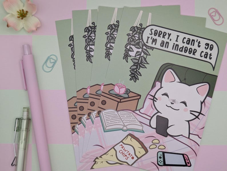 May include: A stack of art prints featuring a cartoon cat in bed with a phone, a book, and snacks. The text reads, "Sorry, I can't go I'm an indoor cat." The prints are on a light green background.