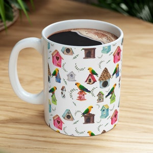 Könnte beinhalten: Eine weiße Keramiktasse mit einem bunten Muster aus Vogelhäusern und Vögeln. Die Tasse ist mit schwarzem Kaffee gefüllt.