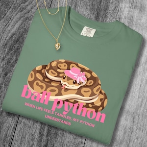 May include: Sage green t-shirt with a graphic of a ball python wearing a pink hat. The text "ball python" is in pink, with the phrase "WHEN LIFE FEELS TANGLED, MY PYTHON UNDERSTANDS" below. A gold necklace with a leaf pendant is draped on the shirt.