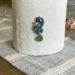 May include: A white, textured cylindrical candle holder with a jeweled seahorse design in shades of blue, green, and gold. The seahorse is affixed to the front of the holder. The candle holder is a decorative item.