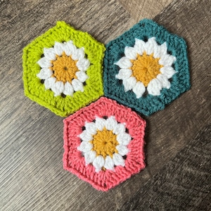 Könnte beinhalten: Drei gehäkelte sechseckige Untersetzer mit einem Gänseblümchen-Design. Die Untersetzer sind grün, türkis und rosa mit weißen und gelben Akzenten.