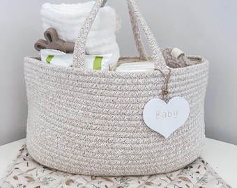 May include: A beige rope basket with handles filled with baby items, including a white washcloth, a brown bunny toy, and a white heart-shaped tag that says "Baby". The basket is sitting on a beige and brown patterned changing mat.