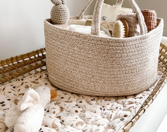 May include: A beige woven storage basket with handles, filled with baby items, including a crocheted bunny toy, a washcloth, and a wooden brush. The basket is sitting on a white and brown patterned changing mat.