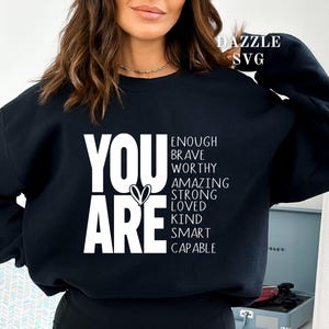 May include: Black sweatshirt with the words "YOU ARE" in large white letters, with a heart symbol. Below, the words "ENOUGH, BRAVE, WORTHY, AMAZING, STRONG, LOVED, KIND, SMART, CAPABLE" are listed. The sweatshirt is a casual style.