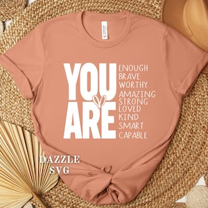 May include: A peach-colored t-shirt with the words "YOU ARE" in large white letters, accompanied by a list of positive affirmations. The shirt is knotted at the bottom and rests on a woven surface.