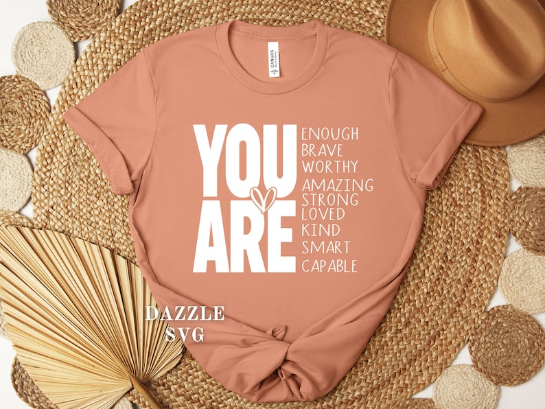 May include: A peach-colored t-shirt with the words "YOU ARE" in large white letters, accompanied by a list of positive affirmations. The shirt is knotted at the bottom and rests on a woven surface.