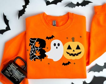 May include: An orange long-sleeve sweatshirt featuring a graphic print that spells BOO. The letter B is black with a white spiderweb, the O is a white ghost, and the second O is a yellow jack-o'-lantern. Small black bats and stars surround the text.