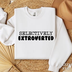 May include: A white sweatshirt with the words "SELECTIVELY EXTROVERTED" in black block letters. The sweatshirt is displayed on a woven surface with a brown hat and decorative elements.