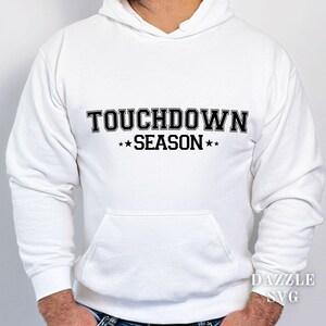 May include: White hoodie featuring the text "TOUCHDOWN SEASON" in black. The text is in a bold, block font, with stars either side of "SEASON". The hoodie has a front pocket and a drawstring hood. The image also includes the text "DAZZLE SVG".