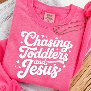 May include: A bright pink crewneck sweatshirt with the text "Chasing Toddlers and Jesus" in white script. The sweatshirt is made of a soft material and is a comfortable fit. A silver necklace is draped over the sweatshirt.