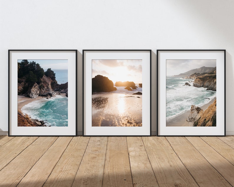 May include: Three framed photographs of a rocky coastline with crashing waves and a sandy beach. The photos are in black frames and are hung on a white wall.