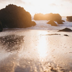 May include: A close-up of a sandy beach with the sun reflecting off the water. The water is calm and clear, and the sand is white and soft. The sun is setting in the background, casting a warm glow over the scene.