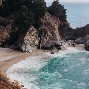 May include: A cascading waterfall flows down a rocky cliffside into a turquoise ocean cove. The beach is sandy with white waves crashing on the shore.
