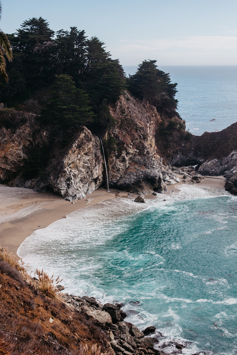 May include: A cascading waterfall flows down a rocky cliffside into a turquoise ocean cove. The beach is sandy with white waves crashing on the shore.