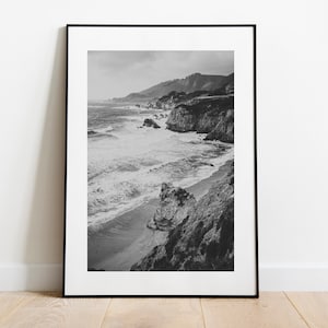 May include: Black and white framed print of a coastal landscape. The photograph captures a rugged coastline with crashing waves, a sandy beach, and distant hills under a cloudy sky. The image is presented in a black frame.