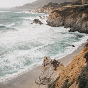 May include: A rocky coastline with a sandy beach and turquoise water crashing against the shore. The cliffs are covered in brown and green vegetation.