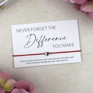 May include: A thin red cord bracelet featuring a small silver-toned heart charm, presented on a white rectangular card with the black printed text: NEVER FORGET THE Difference YOU MAKE. Tie this bracelet around your wrist, close your eyes and make a wish.