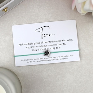 May include: A teal cord bracelet featuring a small silver star charm, mounted on a white display card with the text "Team" and a message about talented people working together to achieve results, plus instructions to make a wish.