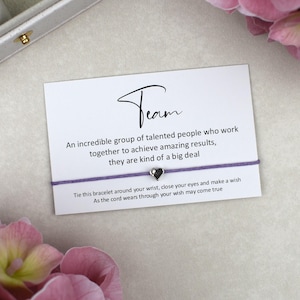 May include: A purple string bracelet with a small silver heart charm attached to a white rectangular card. The card features the text Team, An incredible group of talented people who work together to achieve amazing results, they are kind of a big deal.