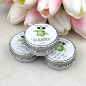 May include: Three small, silver metal tins with white labels featuring a green turtle wearing sunglasses. The text on the label reads "Travel Turtle" and a short poem. Pink and white flowers are in the background.