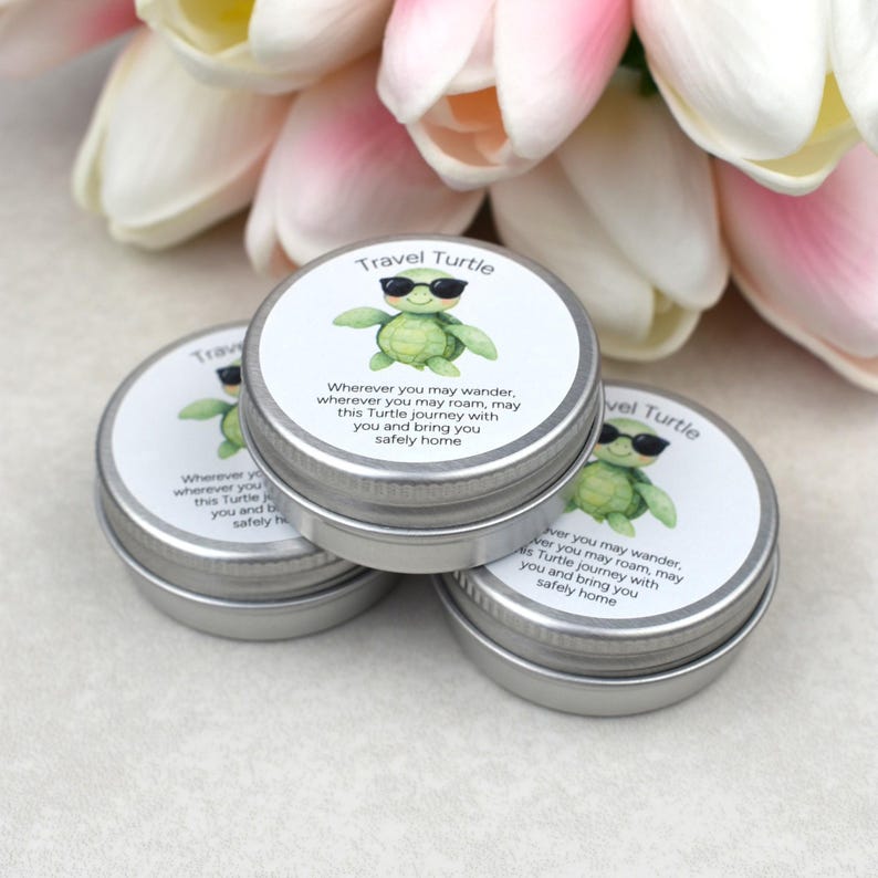 May include: Three small, silver metal tins with white labels featuring a green turtle wearing sunglasses. The text on the label reads "Travel Turtle" and a short poem. Pink and white flowers are in the background.