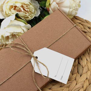 May include: A brown cardboard box wrapped with twine and a blank white gift tag. The tag has the words "To:" and "From:" printed on it. The box is sitting on a woven wicker surface.