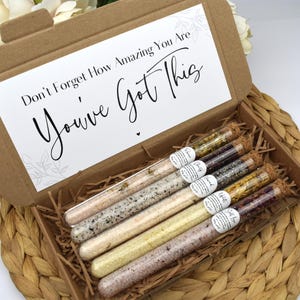 May include: A cardboard box with a white card on top that says "Don't Forget How Amazing You Are You've Got This". The box contains six glass test tubes filled with bath salts and dried flowers. The test tubes are labeled with different scents, including "Rose", "Chamomile", "Lavender", "White Tea", "Salt Love", and "Wild Rose".
