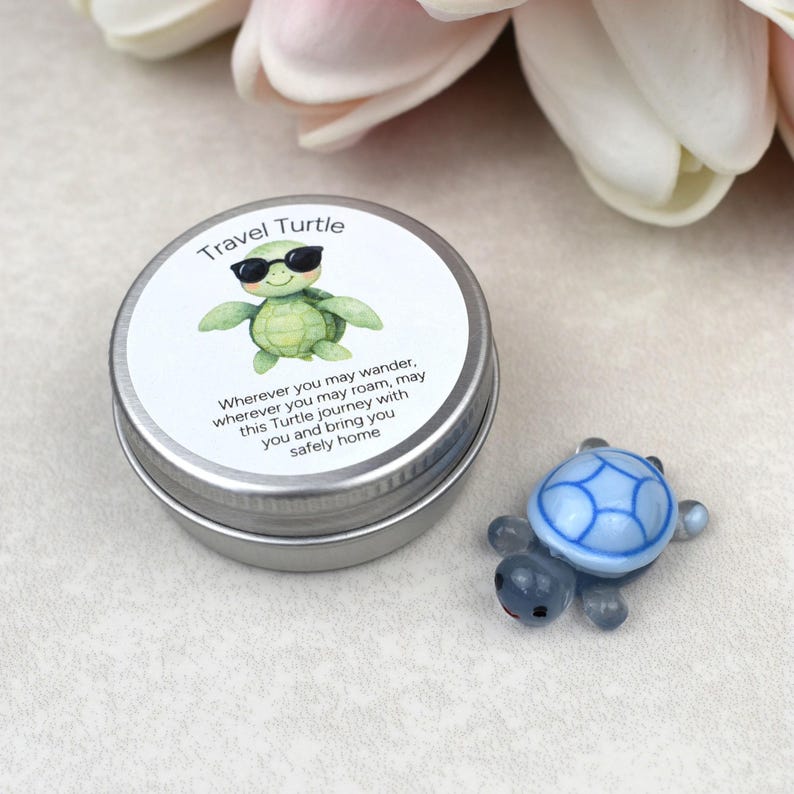 May include: A small, silver tin with a lid featuring a cartoon turtle wearing sunglasses and the words "Travel Turtle." Next to the tin is a small, blue glass turtle figurine. The text on the tin reads, "Wherever you may wander, wherever you may roam, may this Turtle journey with you and bring you safely home."