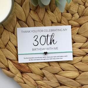 May include: A white card with black text that says "Thank you for celebrating my 30th birthday with me."  A turquoise string bracelet with a silver heart charm is tied to the card. The card is sitting on a woven wicker surface.