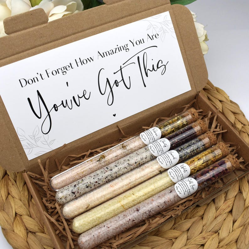 May include: A cardboard box filled with six glass test tubes containing bath salts. The test tubes are labeled with different flower names. The box lid has a white card with the text "Don't Forget How Amazing You Are You've Got This".