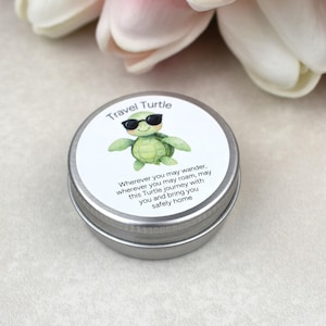 May include: A small, round, silver tin with a white label featuring a green turtle wearing sunglasses. The label reads "Travel Turtle" and includes a travel-themed message. The tin is likely a container for a small gift or keepsake.