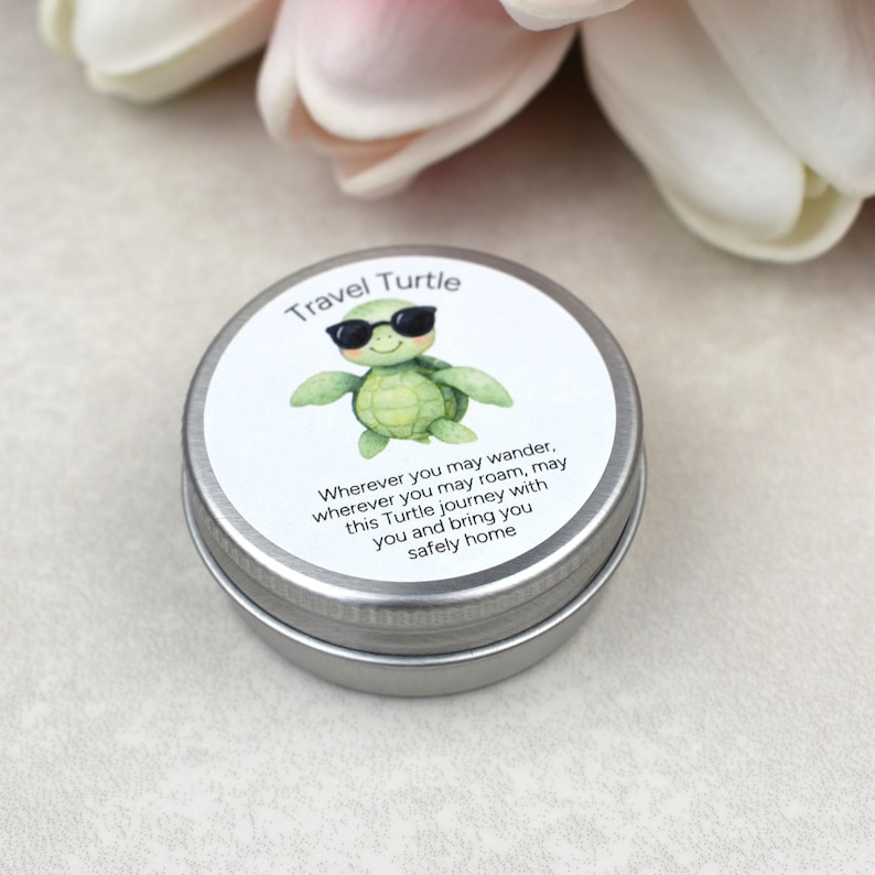 May include: A small, round, silver tin with a white label featuring a green turtle wearing sunglasses. The label reads "Travel Turtle" and includes a travel-themed message. The tin is likely a container for a small gift or keepsake.