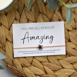 May include: A pink string bracelet with a silver star charm and a white card that reads "YOU ARE ALL KINDS OF Amazing. Tie this bracelet around your wrist, close your eyes and make a wish. As the cord wears through your wish will come true."