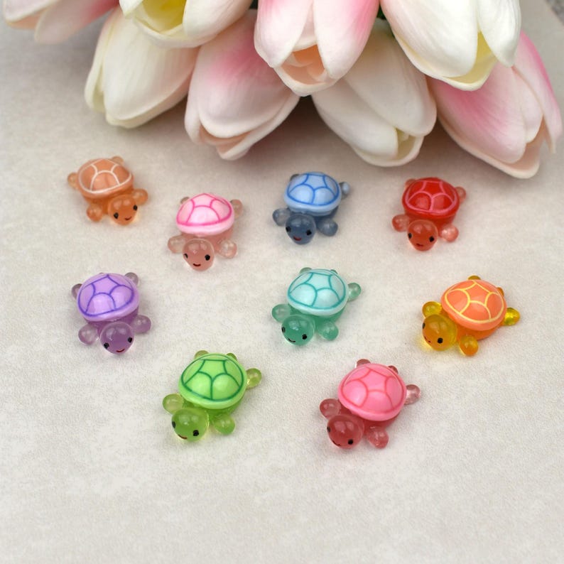 May include: A collection of colorful, miniature turtle figurines in various shades including orange, pink, blue, red, purple, green, and yellow. Each turtle has a smiling face and is made of a translucent material. The turtles are arranged on a white surface.