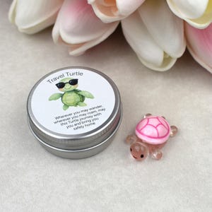 May include: A small, silver tin with a label that reads "Travel Turtle" and features a cartoon turtle wearing sunglasses. Beside it is a small, pink and clear glass turtle figurine.