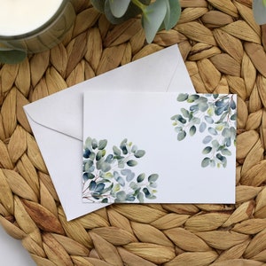 May include: A white card with a watercolour design of green eucalyptus leaves on a woven wicker background.