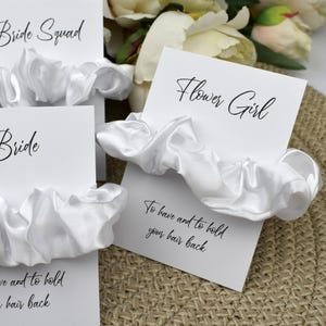 May include: White satin scrunchies with cards that say "Bride Squad", "Bride", "Flower Girl", and the text "To have and to hold your hair back".