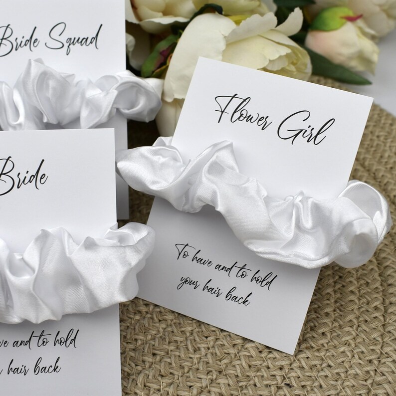 May include: White satin scrunchies with cards that say "Bride Squad", "Bride", "Flower Girl", and the text "To have and to hold your hair back".