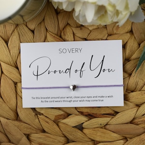 May include: A white card with the text "SO VERY Proud of You" in black script. A purple string bracelet with a silver heart charm is attached to the card. The text on the card reads: "Tie this bracelet around your wrist, close your eyes and make a wish. As the cord wears through your wish may come true."